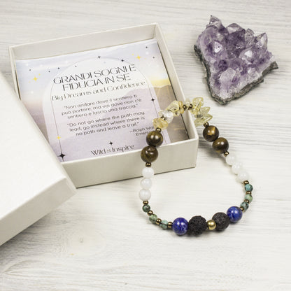 A beaded bracelet with white, dark brown, blue, and black beads is displayed next to its presentation box containing a card that reads ' Big Dreams and Confidence'. A purple amethyst crystal cluster sits in the background on a white wood surface.