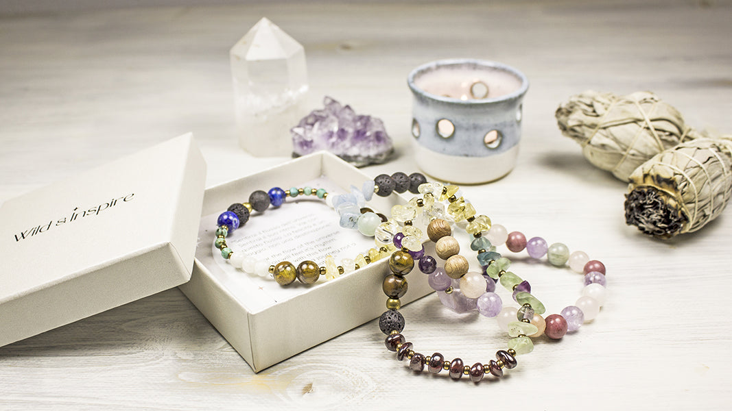 A set of colorful gemstone bead bracelets displayed in an open white gift box, surrounded by crystals, a candle, and a sage stick on a light wooden surface.